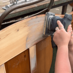 A hand holds a black sander to a brown piece of brown wood, metal bars are above the wood.