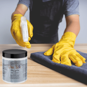 A person with a black apron and grey sleeves, wearing yellow rubber gloves holds a spray bottle pointed to a brown table in one hand and is wiping the table with a blue cloth with the other hand. A jar of Dixie Belle White Lightning is on the tabletop next to the blue rag.