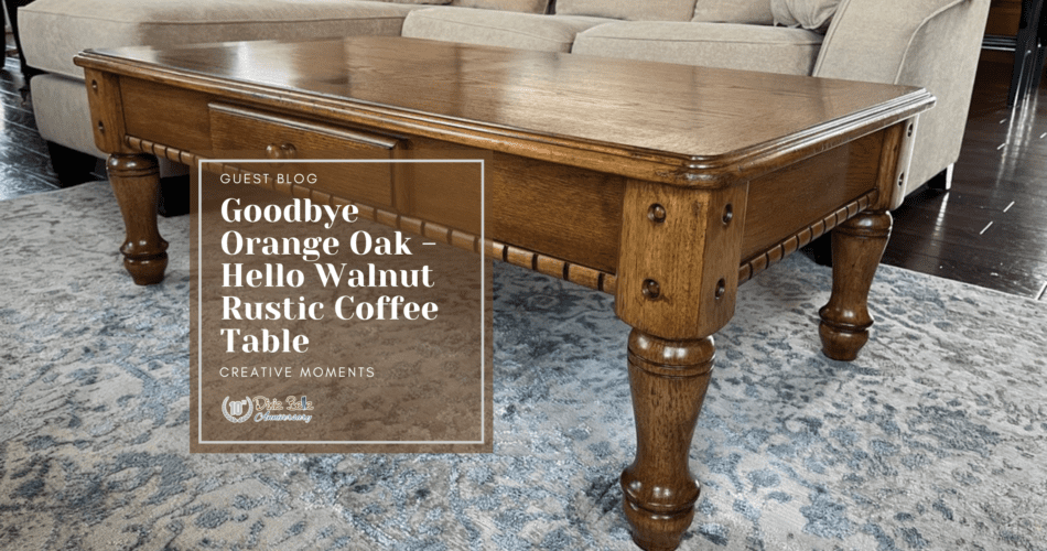 A brown wooden coffee table sitting on a blue and gray rug. A green plant sits on top of the table in a black pot. A tan sofa with a white blanket draped over it is behind the table. In front of the table is a brown box with the words Guest Blog, Goodbye Orange Oak - Hello Walnut Rustic Coffee Table Creative Moments printed in white.