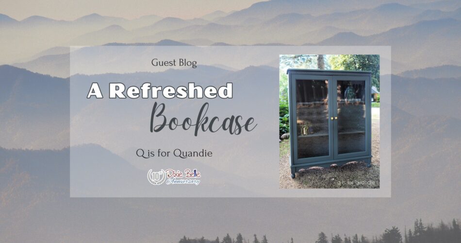 A tall gray painted cabinet with glass doors that have gold knobs sitting on a dirt ground with trees and greenery in the background is to the right of the words Guest Blog Refreshed Bookcase Q is for Quandie. The background is the Great Smoky Mountains.