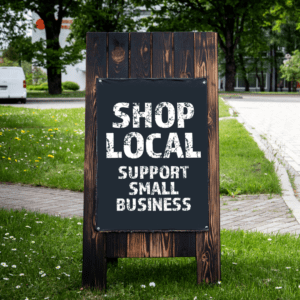 A brown wooden sign is standing on a patch of green grass with a brick sidewalk behind it. The words SHOP LOCAL SUPPORT SMALL BUSINESS are printed on a black board and attached to the sign.