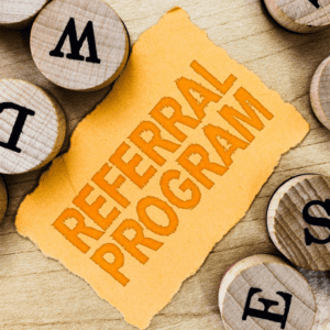 On a yellow sheet of torn paper, the words REFERRAL PROGRAM are printed in orange. The sheet sits on a wooden table, and circular wooden tiles with letters printed on them in black are strewn around the sheet of paper.