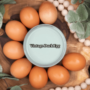 Top view of an open jar of Vintage Duck Egg Chalk Mineral Paint sitting on a circular piece of wood with brown eggs surrounding it and white beads and green foliage around the eggs.