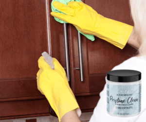 A photo of a person wearing yellow rubber gloves spraying and wiping brown wooden cabinet doors, with a jar of Pristine Clean in the lower right corner.