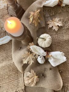 a brown painted wooden table with burlap, a white painted pumpkin, white painted gourd and white napkin, a brown painted candlestick holding a white candle and leaves strewn about.