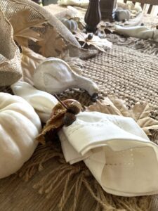 A white napkin tied with a brown piece of cloth sits on a piece of burlap on a brown wooden table. A white painted gourd, a white painted pumpkin and leaves surround the napkin