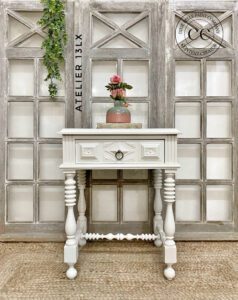 "After" picture of a white painted nightstand sitting on a brown woven rug in front of tan painted window panes in front of a white wall