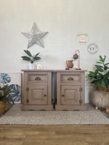 A pair of nightstands painted with Cobblestone Chalk Mineral Paint sitting on a brown rug in front of a white painted wall. Green plants are on the floor to the left and right of the nightstands and knick knacks sit on top of them.