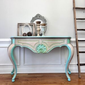 A hallway table stained with Fireside Ash Voodoo Gel Stain, highlighted with Mermaid Tail chalk Mineral Paint sits on a brown wooden floor in front of a white wall. A wooden ladder is to the right of the table and there are candlesticks and photo frames on top of the table.