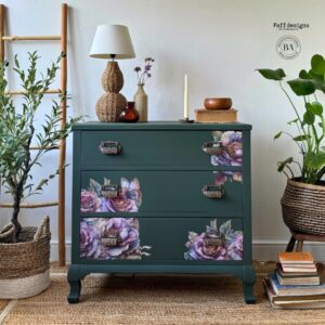 A green painted dresser with the My Favorite Bouquet transfer applied in various spots on the drawers; sitting on a brown rug in front of a white wall; a wooden ladder and a green tree in a basket sit to the left of the dresser; a green plant in a basket and a pile of books sit on the floor to the right of the dresser; a lamp and several different knick knacks sit on top of th edresser.