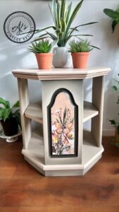 Octagonal table that is painted with Buttercream Chalk Mineral Paint and has the Savi's Bouquet transfer applied to the front center section, sitting on a brown wooden floor in front of a white wall. 3 potted plants sit on top of the table and plants are on the floor on both sides of the table.