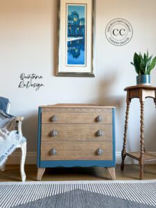 A dresser with blue painted frame and Bleached Cypress stained drawers sitting on a wooden floor, in front of a white wall; a gray and white rug in front of the dresser, a chair is to the left and a plant on a wooden stand is to the right of the dresser; a blue and white picture are hung on the wall above the dresser