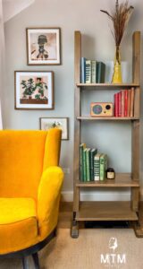 A shelving unit stained with Whiskey Barrel Voodoo Gel Stain has books and other knick knacks on each shelf sitting on a brown wooden floor, with a yellow chair to the left of it. Photos hang on the wall.