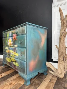 Dresser painted with a blend of various Chalk Mineral Paint colors that has the Lily Pads decoupage paper applied to the front of it sitting on a brown wooden floor, in front of a black painted wall