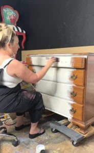 Tracey sitting on a stool applying white paint to the front of the brown wooden dresser