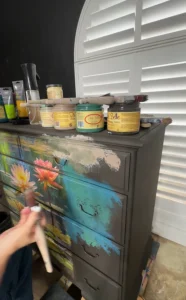 Brown painted dresser with Lily Pads decoupage and various colors of paint being blended on the front of the dresser. Several open jars of Dixie Belle Chalk Mineral Paint and paint brushes sit on top of the dresser.