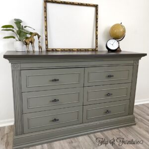 A gray painted dresser with black antiquing sits on a brown wooden floor in front of a white painted wall. A picture frame, green potted plant, globe, clock and bronze horse statue sit on top of the dresser.