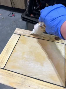 A hand holding a bottle of Voodoo Gel Stain, applying it to the top of a wooden table