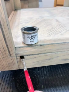 An open container of Black Gilding Wax sits on a stained end table, with a small paintbrush applying gilding wax to the ridge