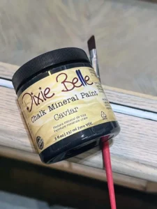 A jar of Caviar Chalk Mineral Paint and a small paint brush sits on top of a wooden end table.