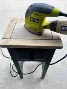 Small black end table with raw wood tabletop being sanded with green and black Ryobi sander